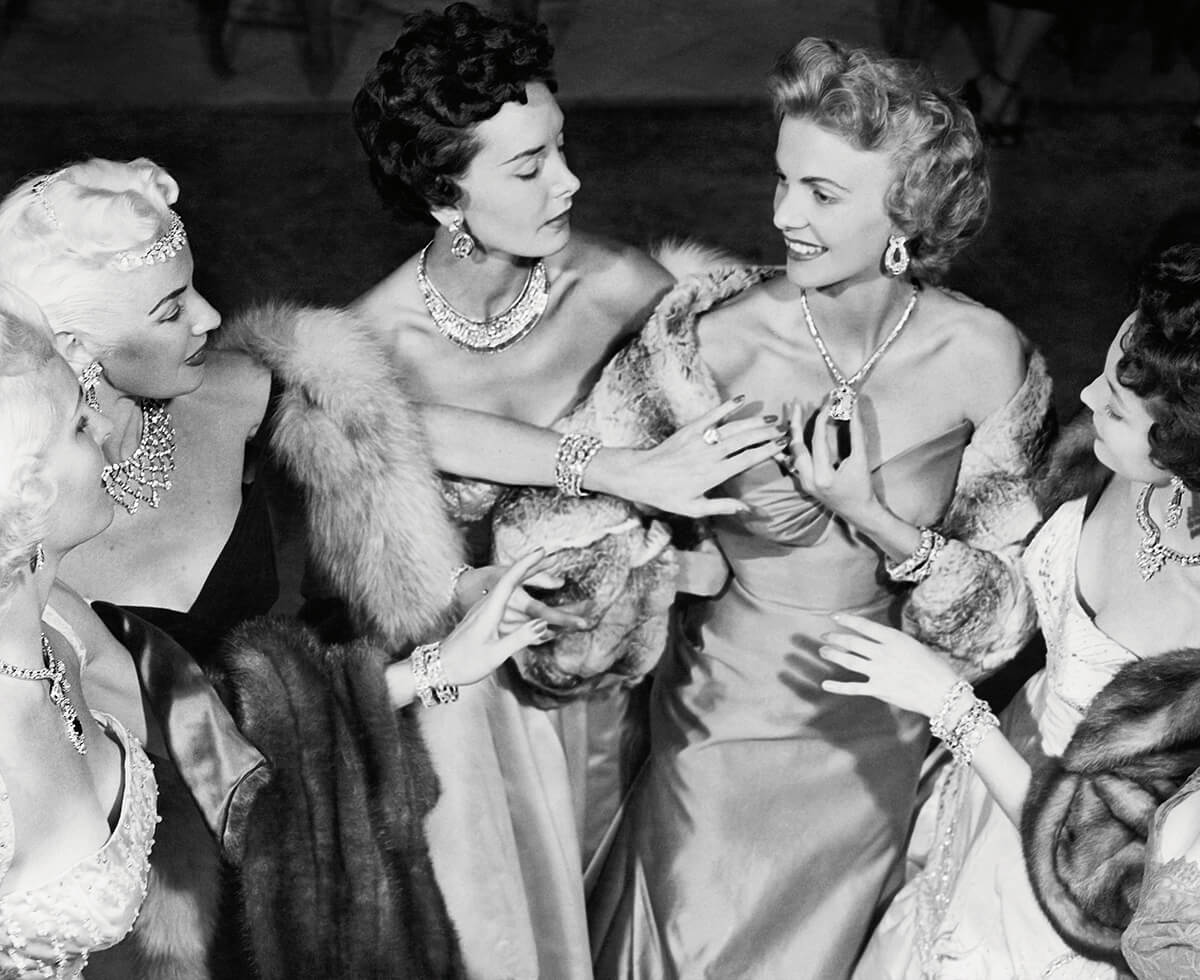 A black and white image shows a group of women adorned in jewelry admiring one another's magnificent necklaces and rings at Harry Winston's Court Of Jewels